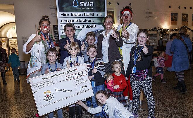 Kinder stehen mit Spendenscheck neben zwei Mitgliedern der KlinikClowns. Weiterverlinkung auf swa Kids Aktionsseite
